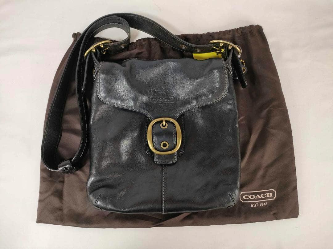 Front view of the Coach Bleecker Legacy flap crossbody bag (black) with buckle, shown on a brown Coach dust bag.