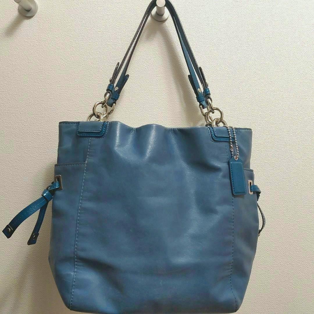 Blue COACH leather shoulder bag hanging from a hook against a light textured wall.