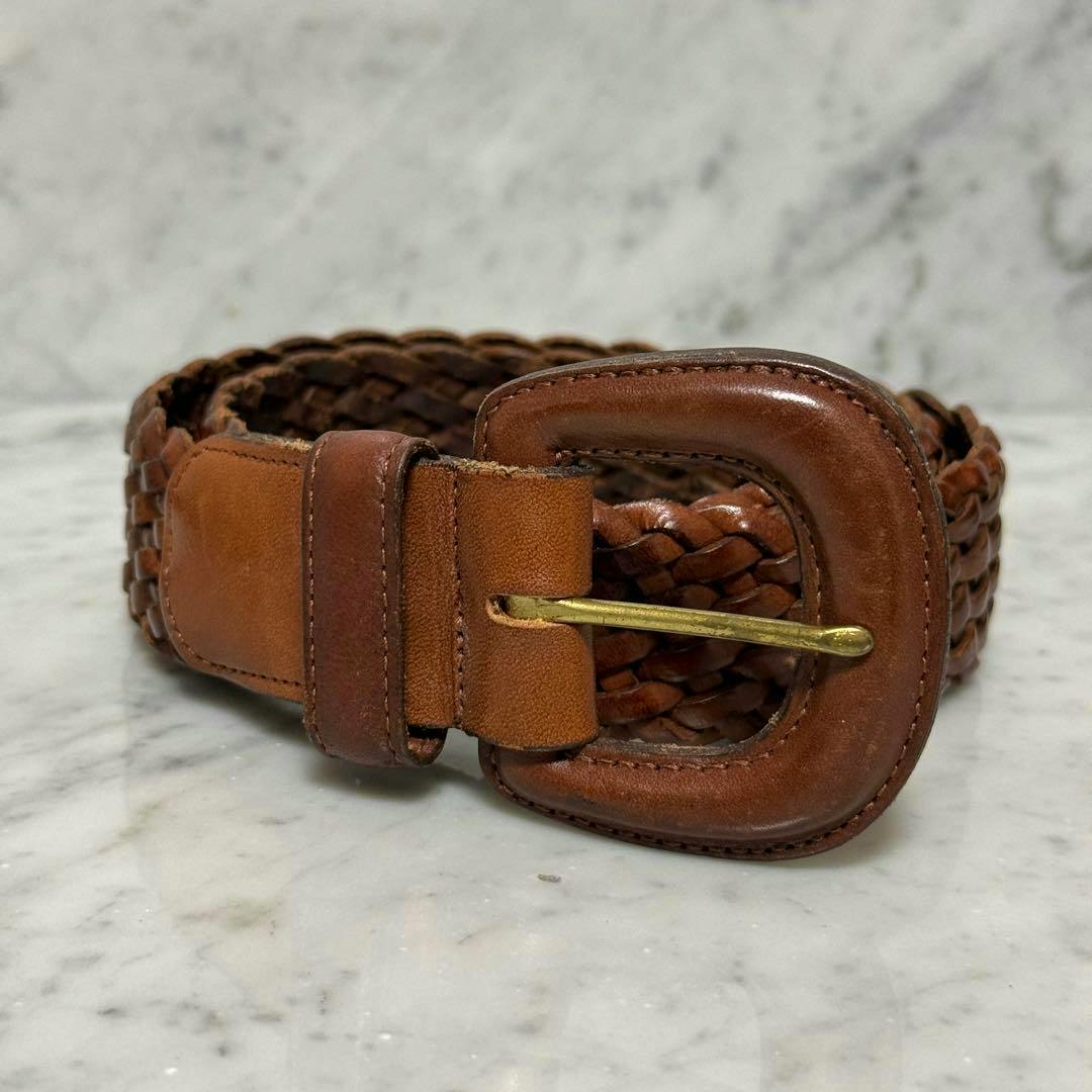Brown braided cow leather belt with a rounded leather-covered buckle and brass pin on a marble surface.