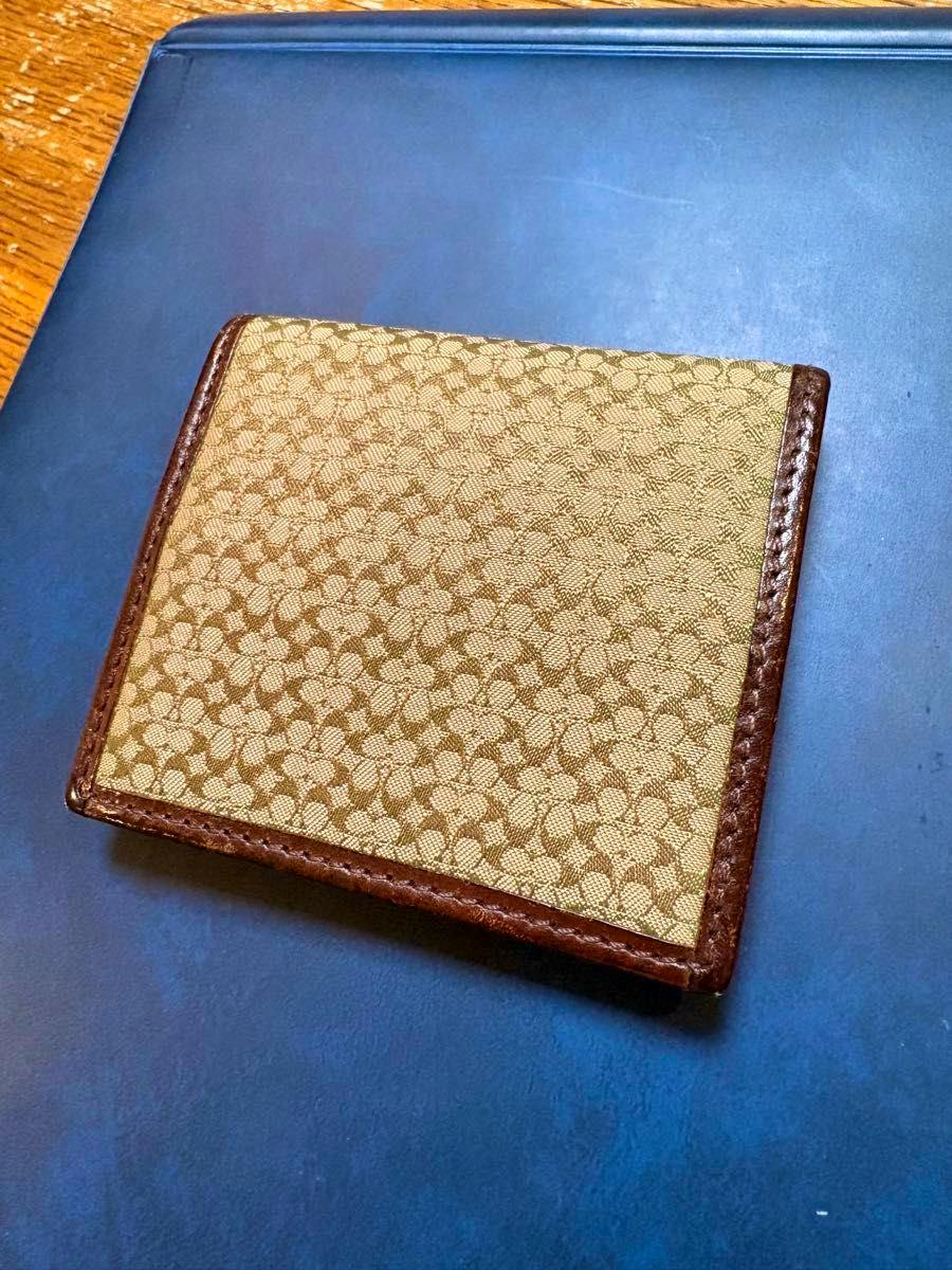 Closed brown COACH coin case in canvas with signature monogram and brown leather trim on a dark surface.