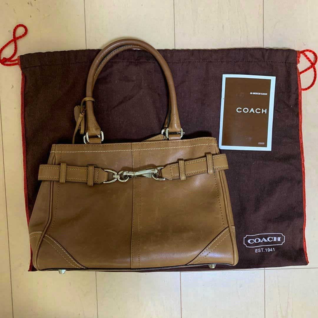 Brown COACH leather handbag placed on a dark Coach dust bag with a COACH booklet beside it.