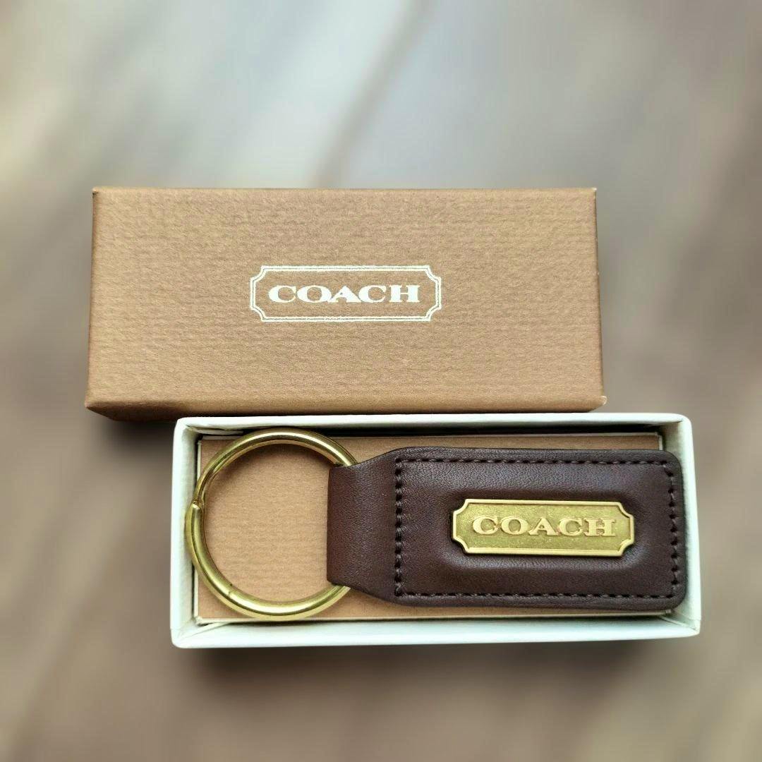 Brown COACH leather keychain with a gold-tone key ring and a COACH metal nameplate displayed in its box.