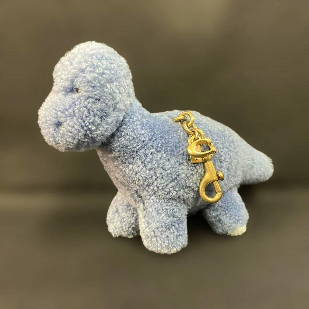 Blue shearling dinosaur-shaped COACH bag charm with gold-tone lobster clasp on a dark background.