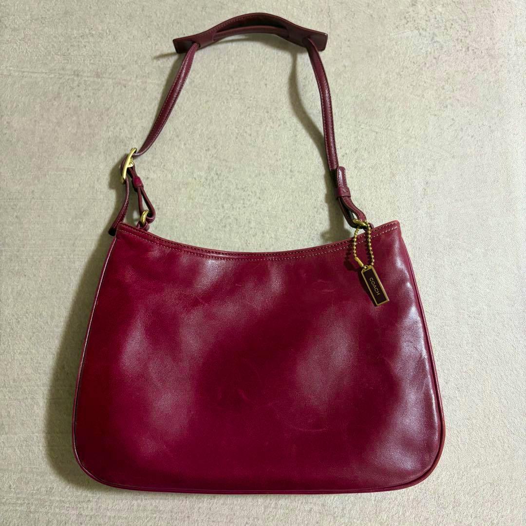 Burgundy/bordeaux leather Coach pouch handbag shown flat with shoulder strap and a Coach hang tag on a light background.
