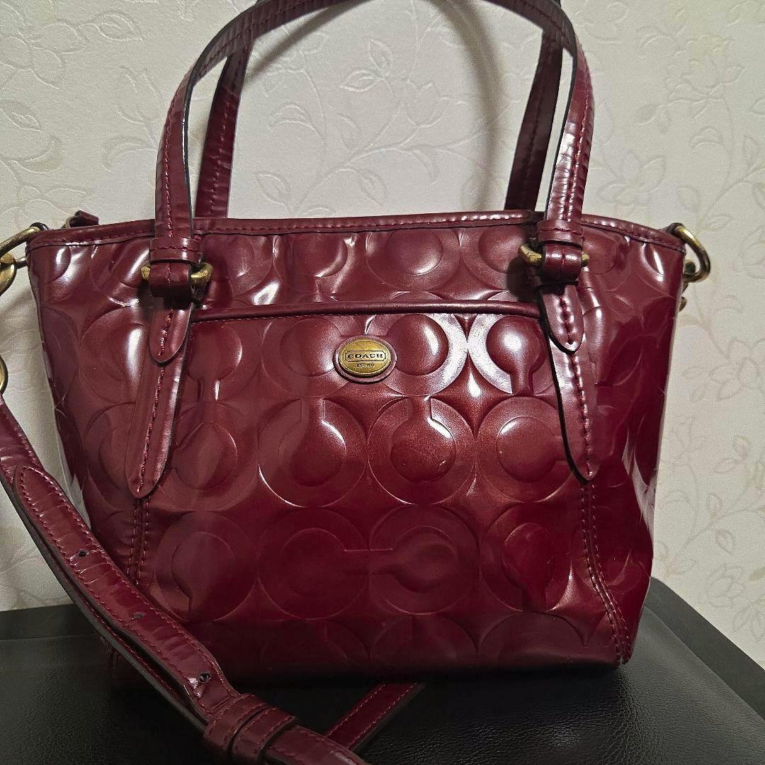 A burgundy patent leather Coach shoulder bag with embossed C pattern and a long strap against a light patterned wall.