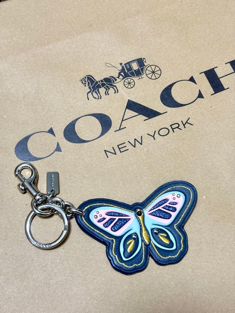Leather butterfly bag charm keyring with metal clasp and logo tag displayed on a COACH New York paper background.