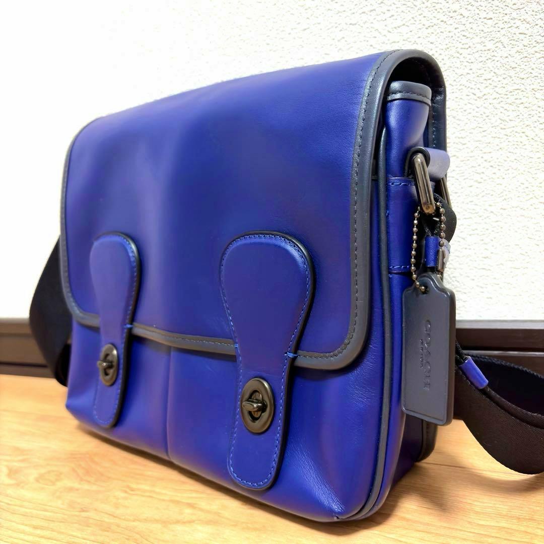 Blue leather COACH map-style shoulder bag shown at an angle on a wooden surface, showing the front flap, turn-lock closures, side strap, and hang tag.