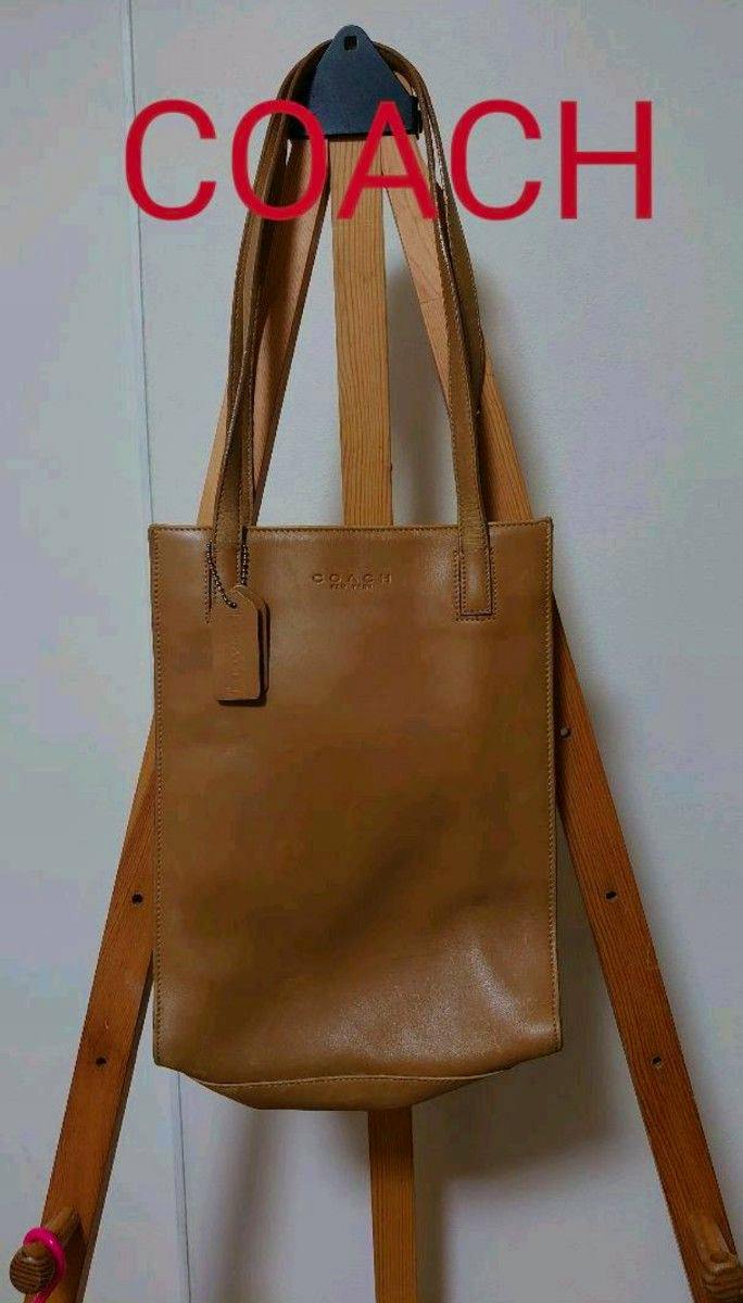 Camel-colored Coach cow leather tote bag hanging on a wooden stand, front view with leather hang tag and red "COACH" text at the top.