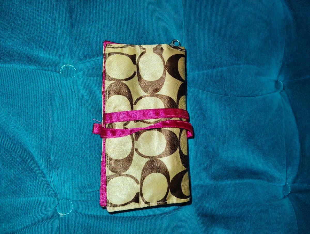 Closed COACH accessory pouch with brown 'C' pattern and pink ribbon tie on a teal background.