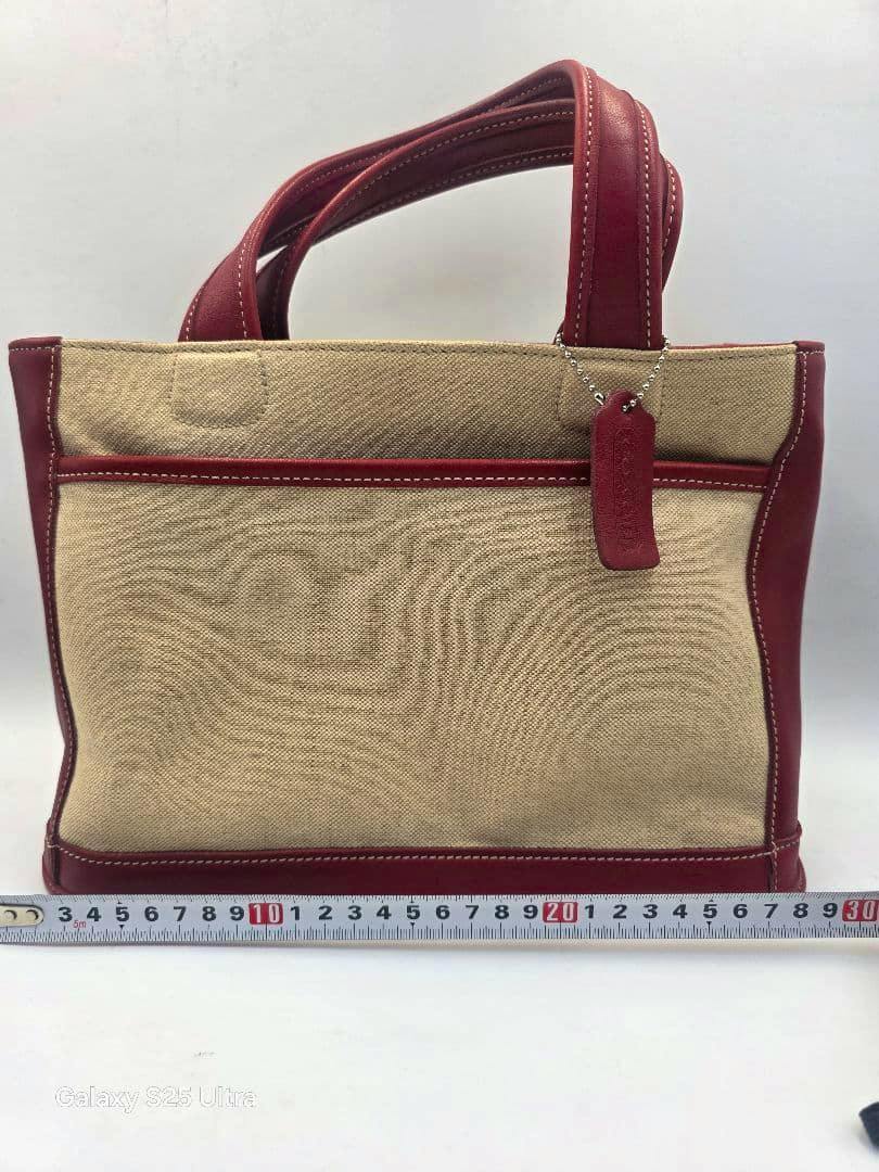 Front view of the COACH canvas and red leather handbag with a leather tag and a measuring tape at the base.