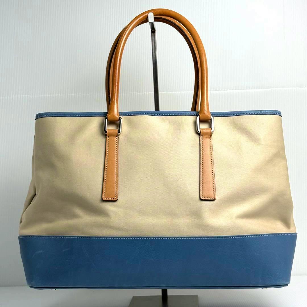 Front view of a COACH beige canvas tote bag with a blue leather base and tan leather handles.