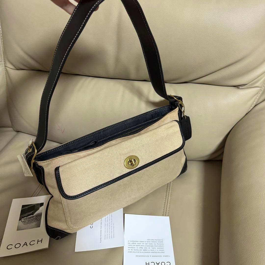 Beige Coach canvas shoulder bag with black leather trim and a gold turn-lock, photographed on a beige sofa with Coach cards nearby.