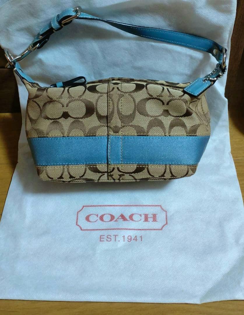 Front view of a Coach canvas bag with brown signature 'C' pattern and a blue fabric/leather trim and strap placed on a white Coach dust bag.