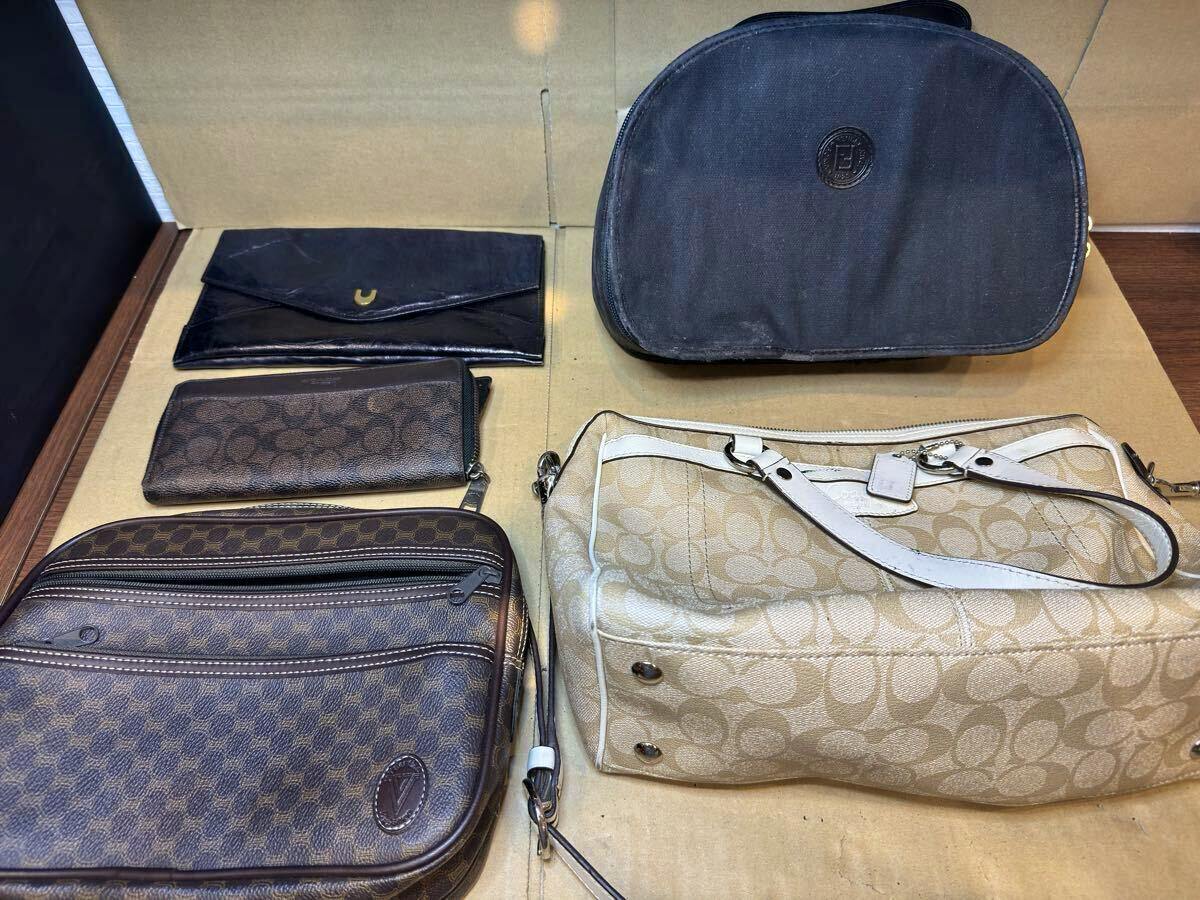 Five bags and wallets arranged on cardboard, including a beige Coach signature shoulder bag, a brown patterned crossbody, a black pouch, a brown zip wallet, and a dark vanity bag.