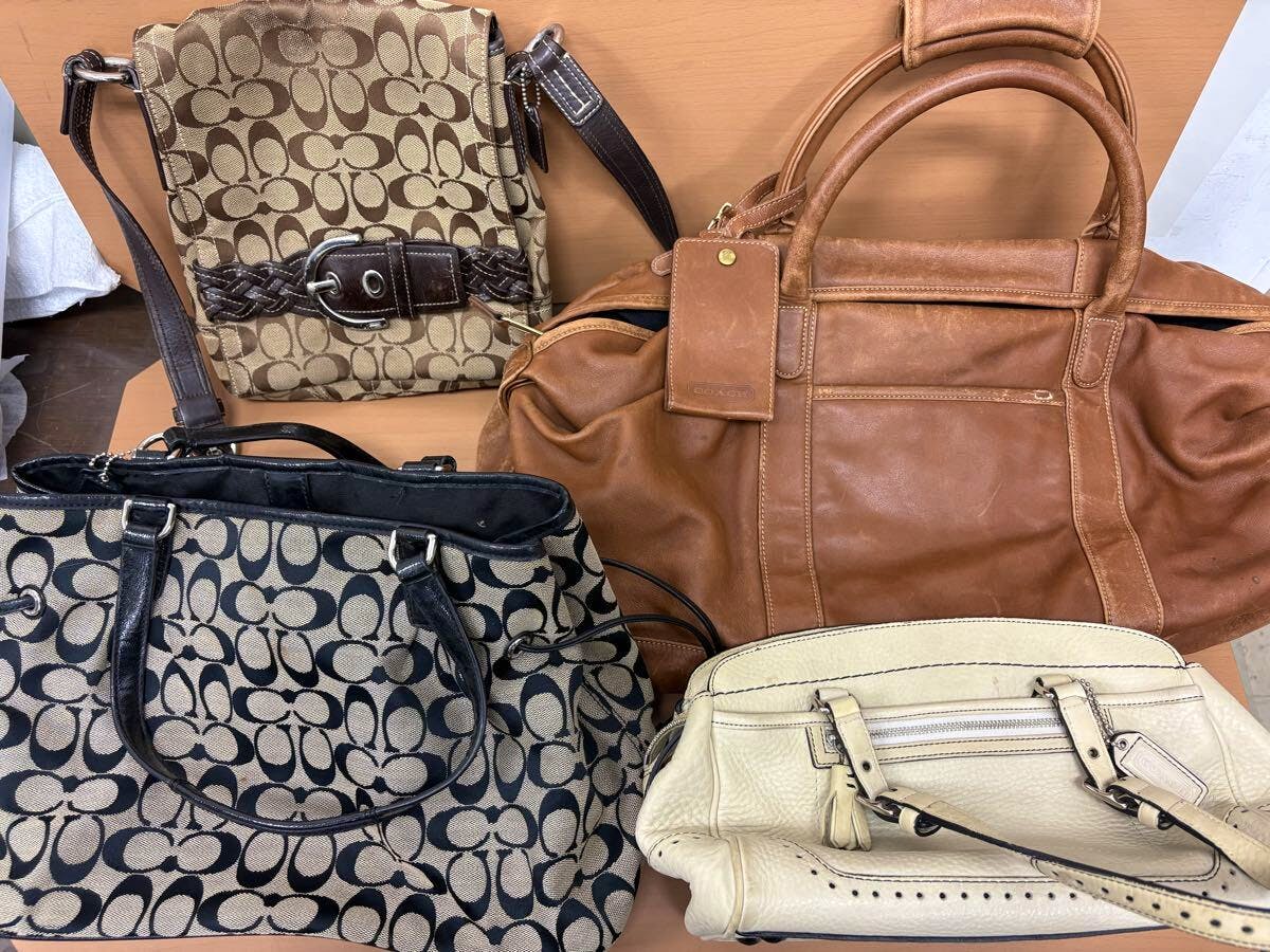 Four Coach bags — a brown leather duffel, two signature canvas bags, and a cream leather handbag — arranged on a surface.