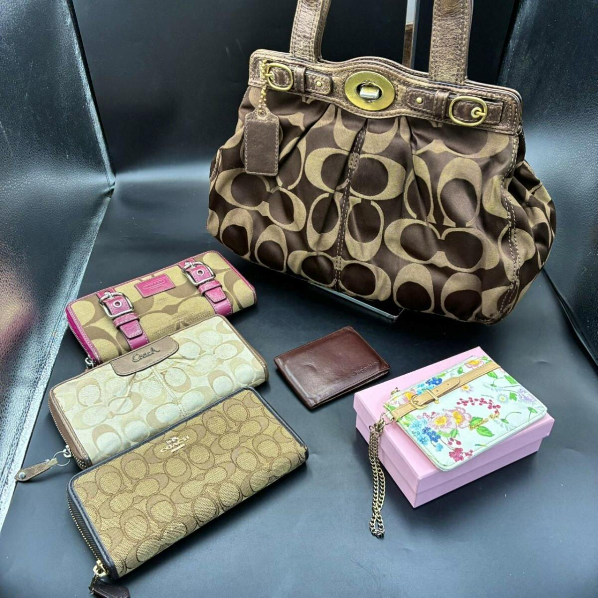 Brown Coach canvas bag with signature 'C' pattern shown with several Coach wallets and a floral card case on a black surface.