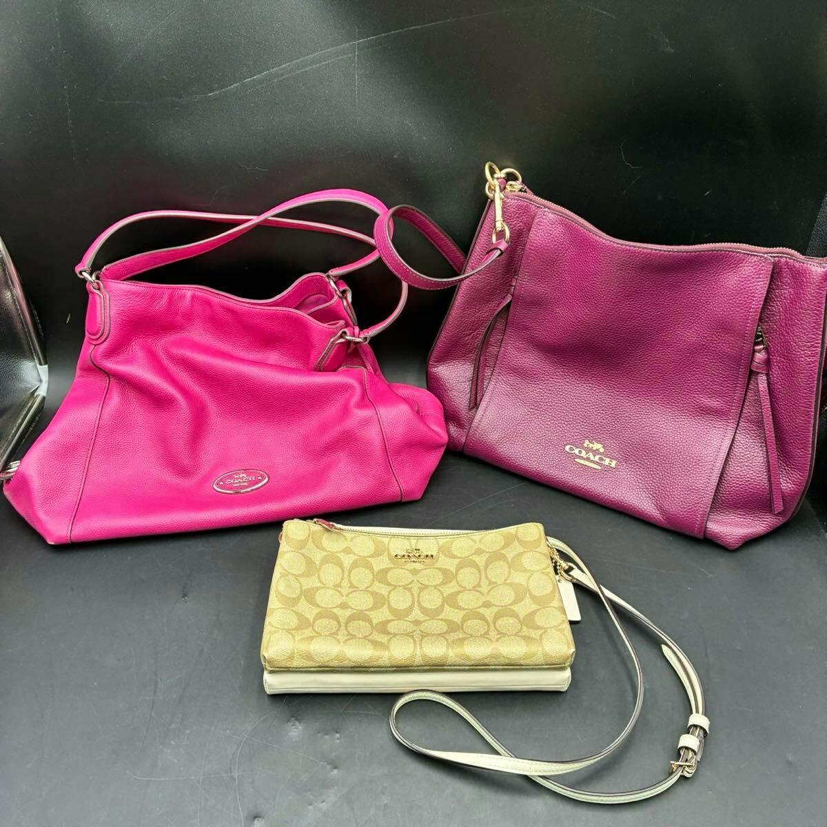 Three Coach bags—a bright pink shoulder bag, a purple tote, and a beige monogram wristlet—placed on a black surface.