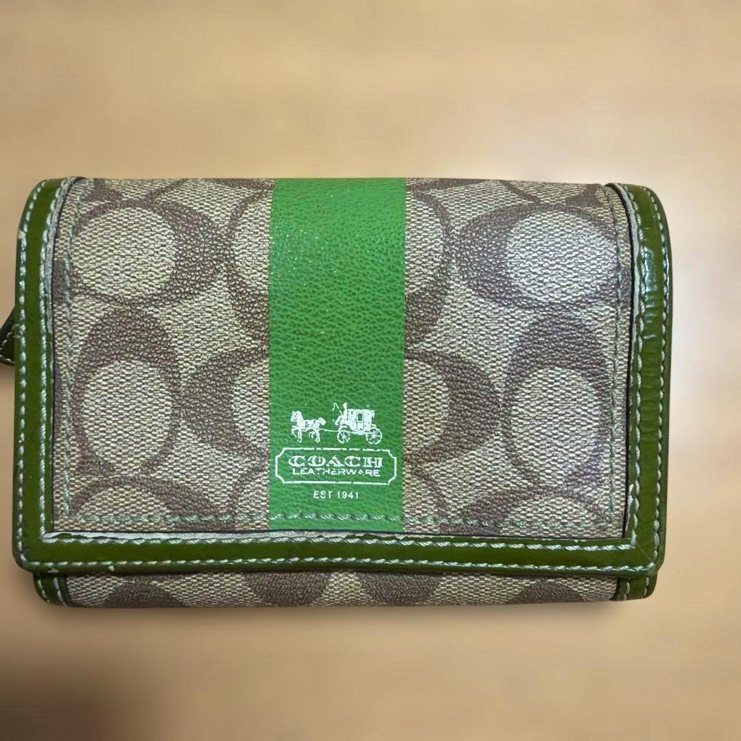 Front of a Coach canvas two-fold wallet showing the signature "C" pattern, a central green vertical stripe, and the Coach logo.