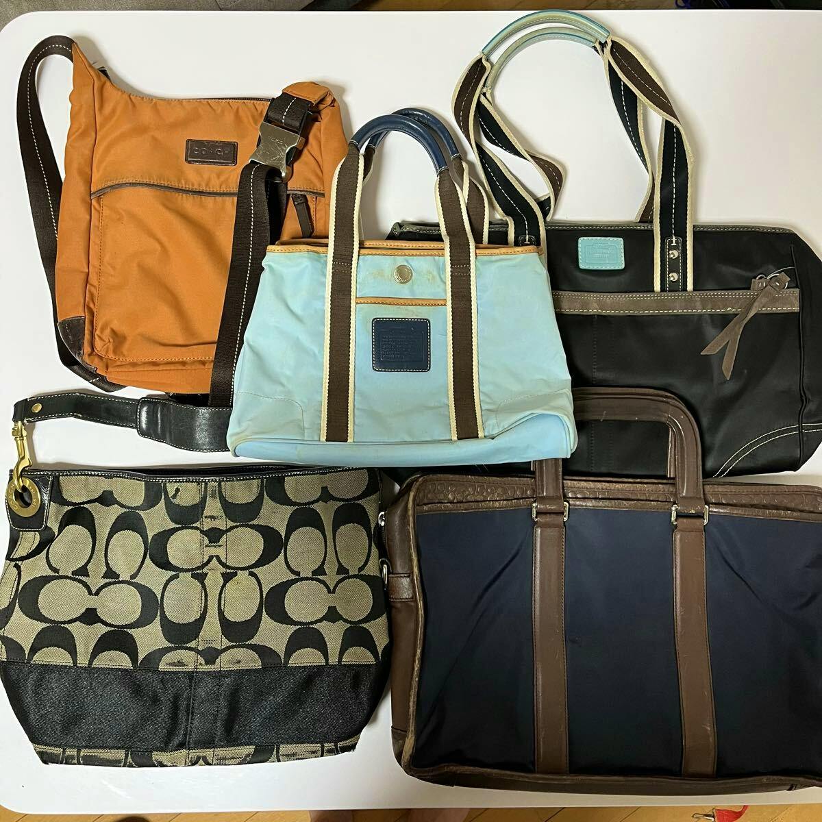 Five COACH canvas and leather handbags and shoulder bags arranged flat on a white surface.