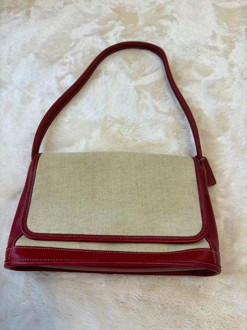 Beige canvas shoulder bag with red leather trim and a single red shoulder strap, shown lying flat.