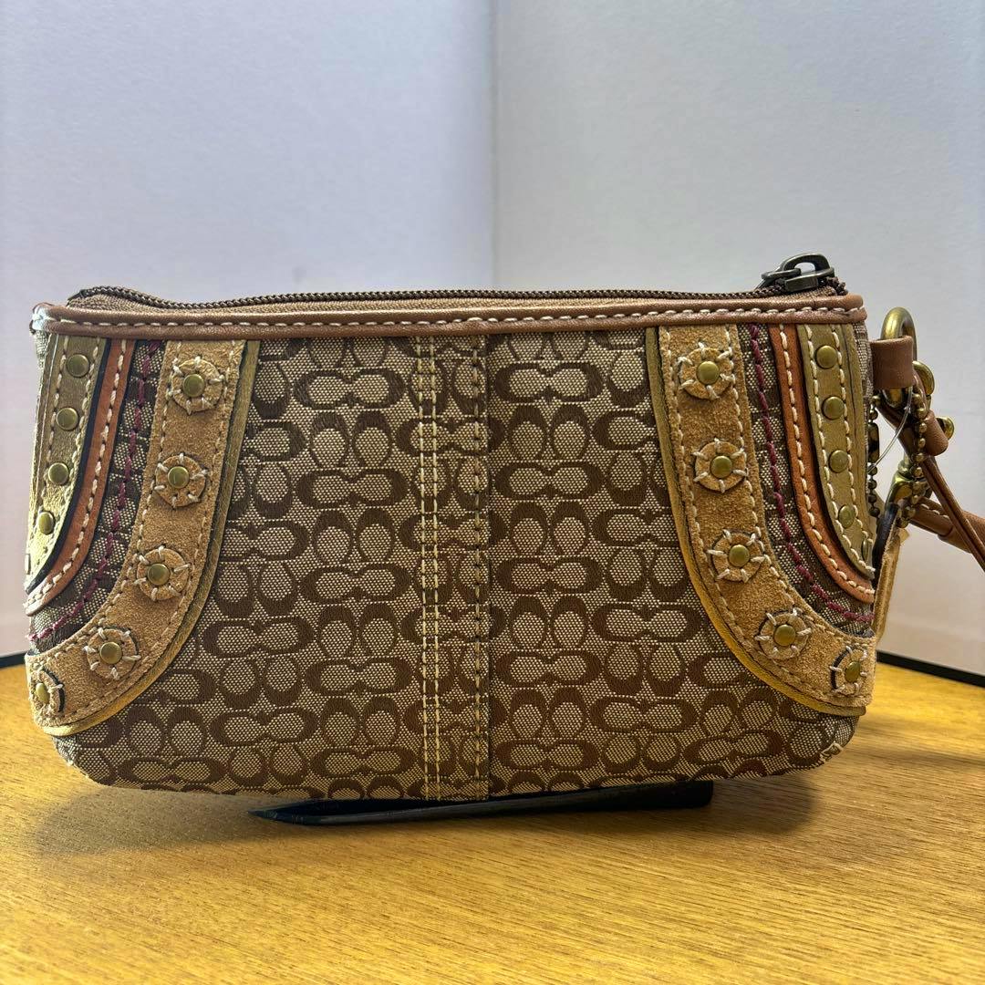 Brown Coach canvas pouch with signature 'C' pattern and studded suede trim shown on a wooden surface.