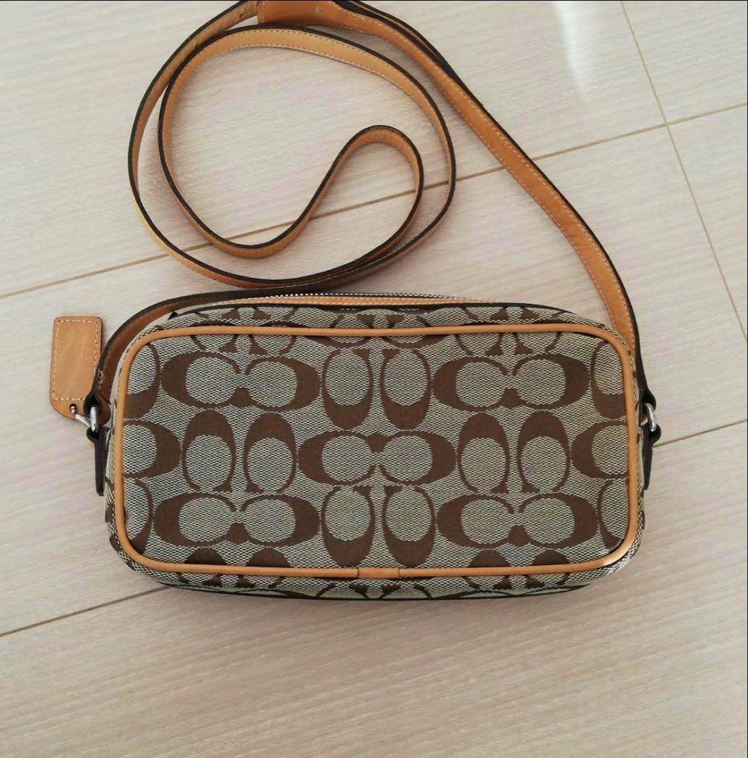 Top-down view of a brown Coach canvas shoulder bag with signature C pattern, tan leather trim, and a long strap on a light floor.