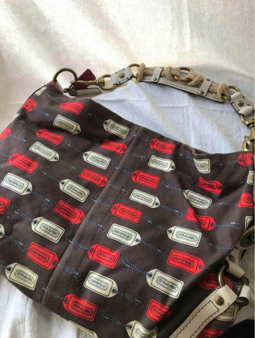 Brown canvas Coach shoulder bag with red and white tag-print pattern and a white leather-and-rope strap shown laid flat.