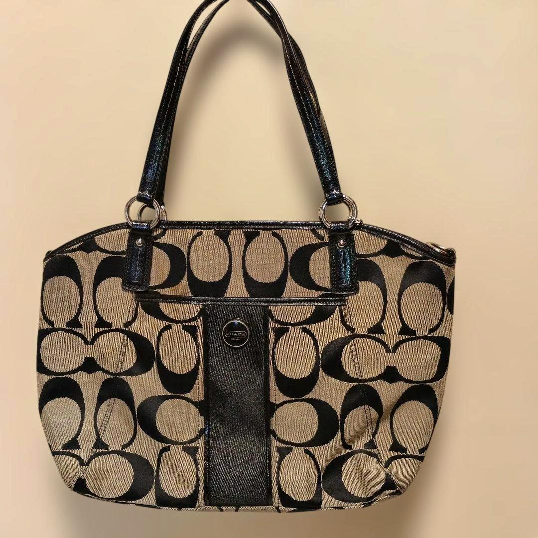 Front view of a beige and black Coach canvas shoulder bag with signature 'C' pattern and black handles.