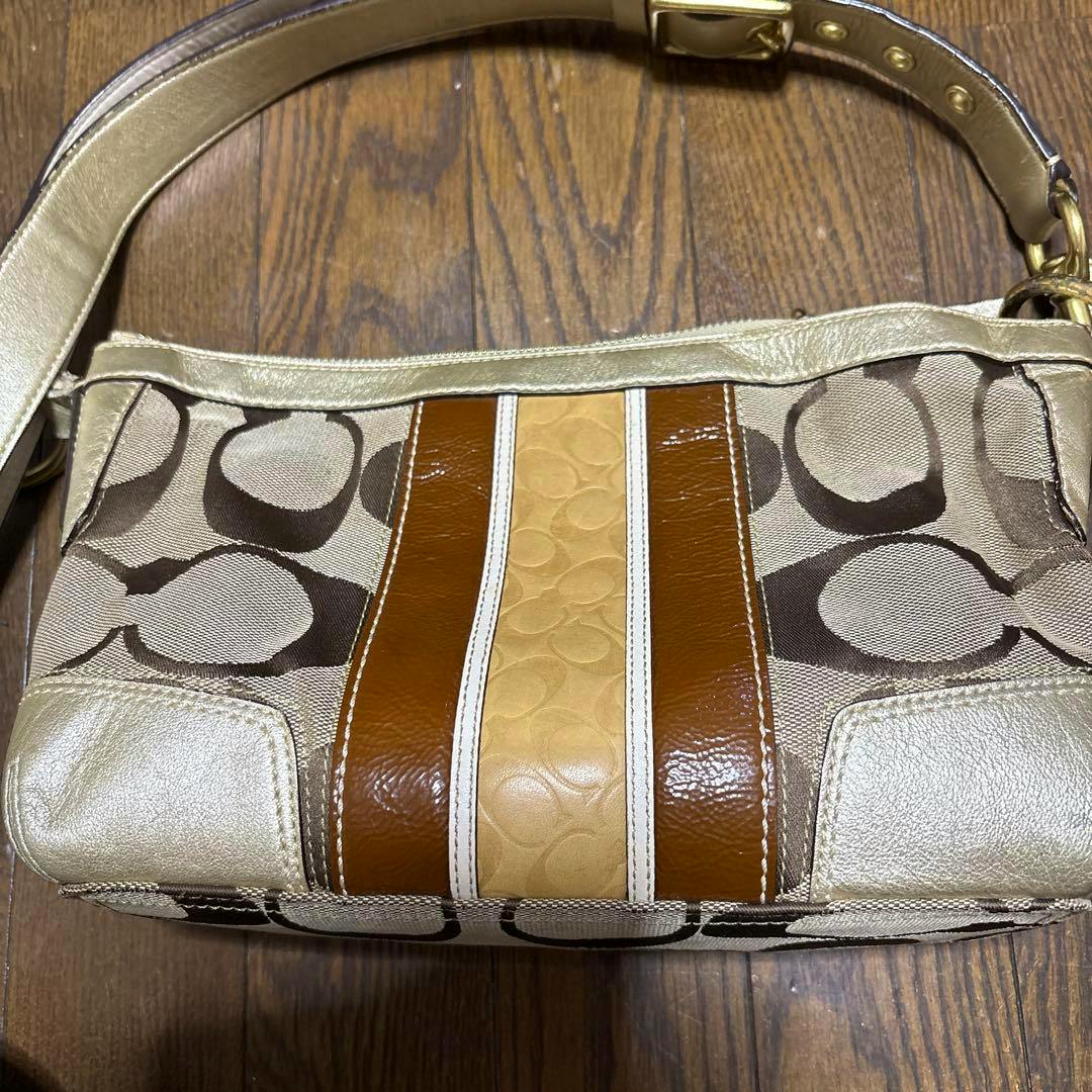 Coach canvas shoulder bag with signature 'C' pattern, center leather stripes, and metallic trim laid on a wooden floor.