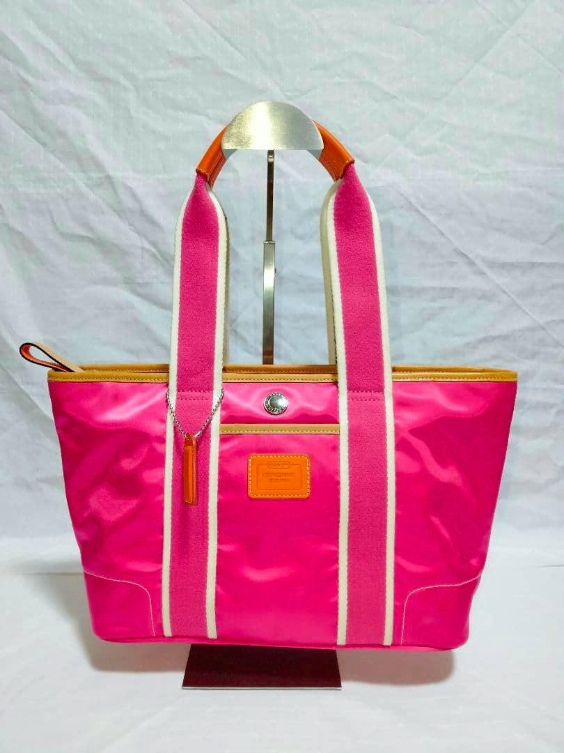 Front view of a pink Coach shoulder/tote bag with wide pink-and-white straps and orange trim displayed on a stand.