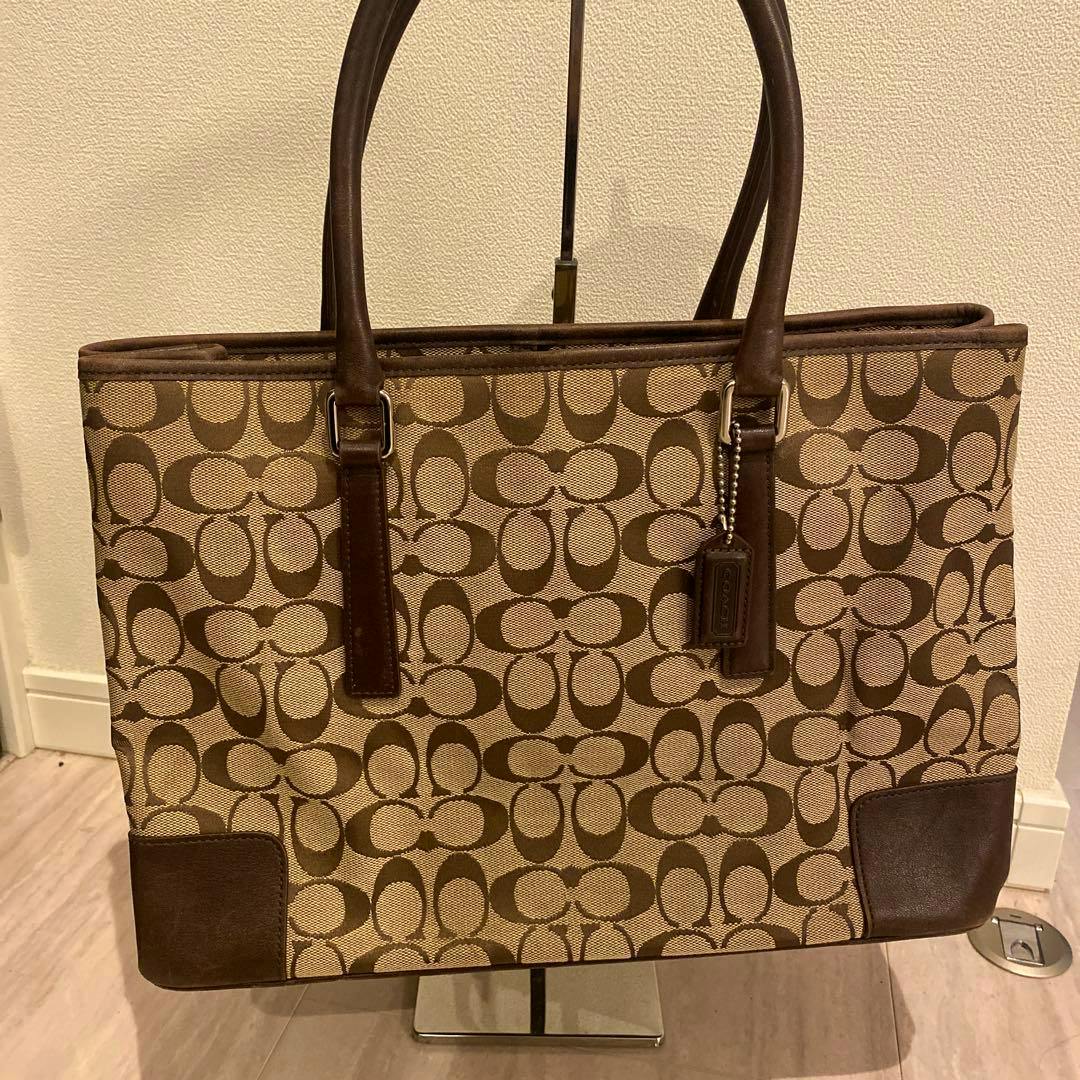 Front view of a beige and brown Coach canvas tote bag showing the signature C pattern and brown leather trim and handles.