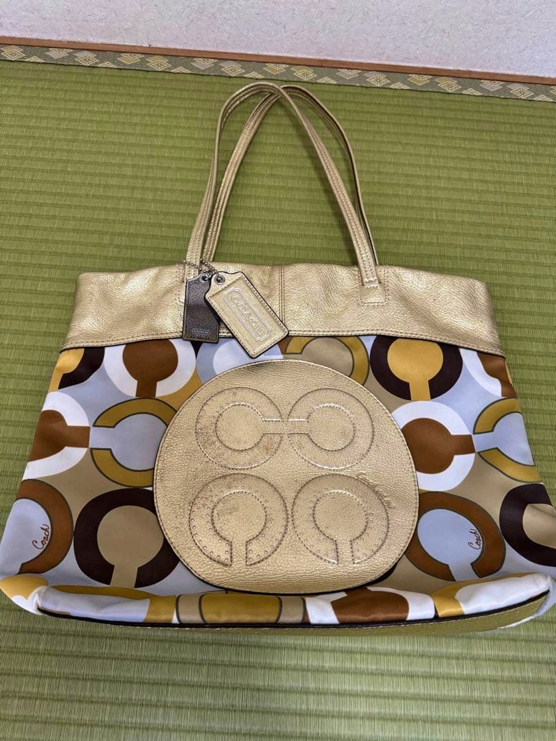 Gold-tone COACH tote bag with patterned canvas body, circular leather emblem, top handles, and Coach hang tag laid flat on a mat.