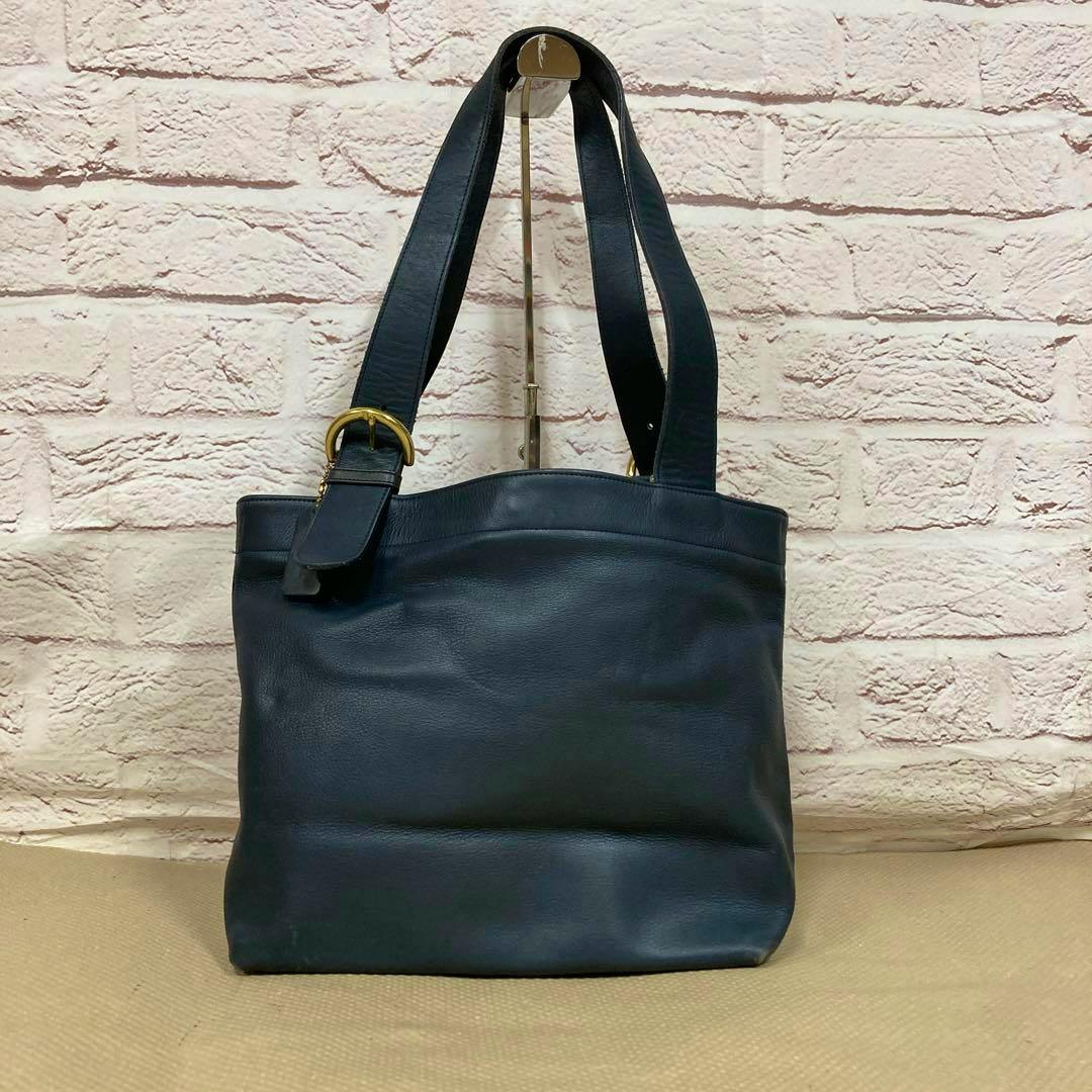 COACH navy tote bag photographed front-on with its double shoulder straps visible against a brick backdrop.