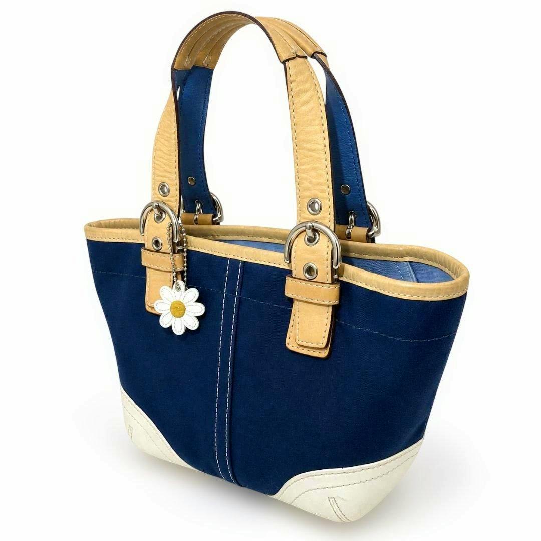 Navy and tan bicolor Coach canvas tote bag with white bottom accents and a daisy flower charm shown at a three-quarter angle.