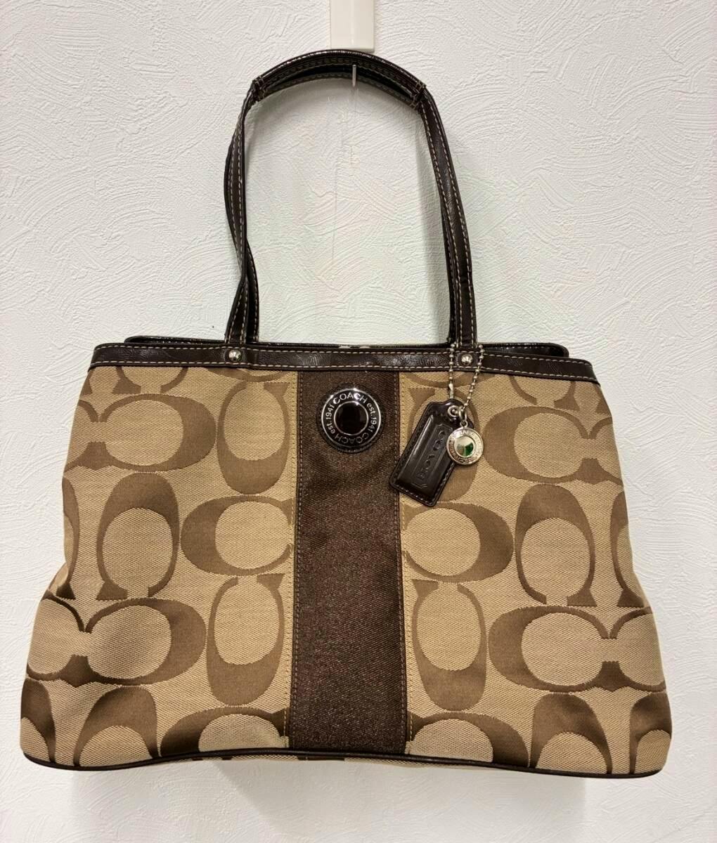 Front view of a brown COACH canvas tote bag with signature 'C' pattern, a dark leather center panel, and hanging Coach tags.