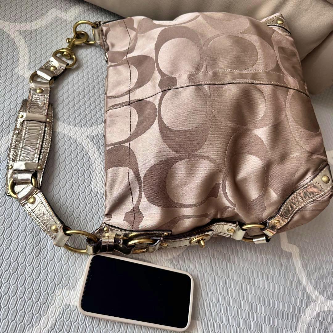 Beige Coach signature canvas Carly Hobo shoulder bag with metallic strap, photographed flat with a smartphone nearby.