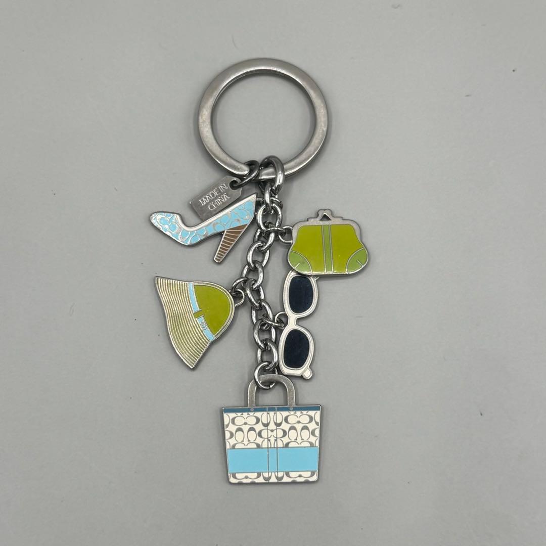 A Coach casual style keychain with metal ring and several colored enamel charms including a tote, sunglasses, hat, shoe, and small purse on a gray background.