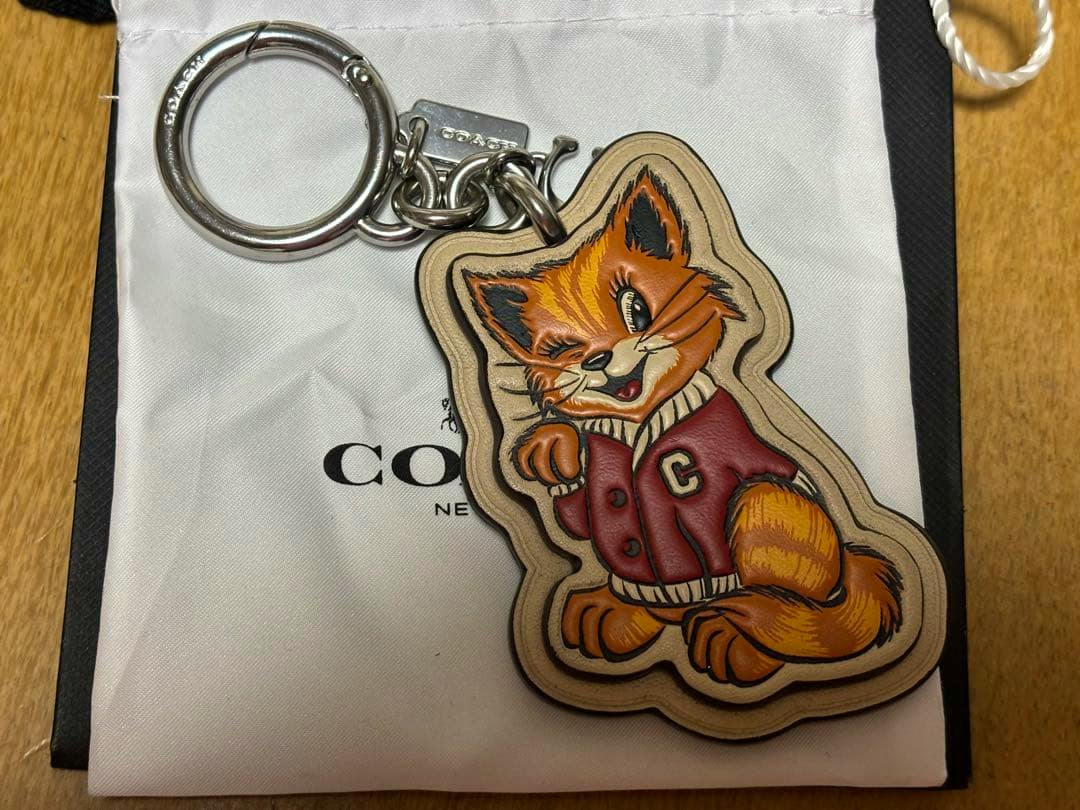 Cow leather keychain bag charm shaped like an orange winking fox wearing a red jacket attached to a metal key ring, shown on a Coach pouch.