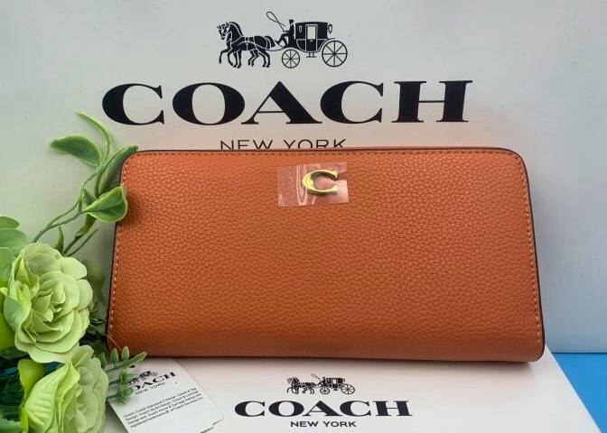 Orange Coach long leather wallet front with a small gold 'C' emblem, placed in front of a COACH New York box and tag.