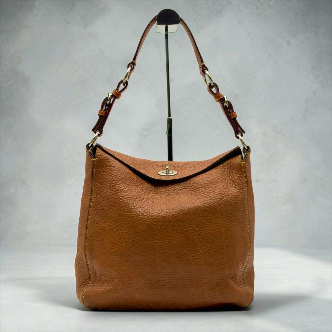 Brown pebbled leather Coach Chelsea shoulder bag with a top flap and turn-lock closure displayed on a stand.