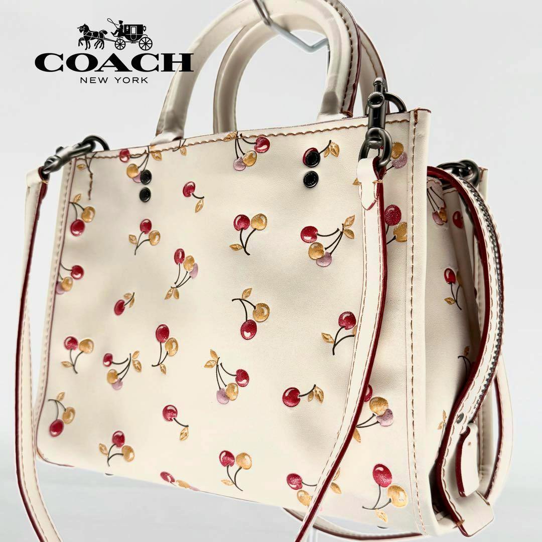 White COACH leather 2-way handbag with small red and gold cherry print and top handles shown on a white background.
