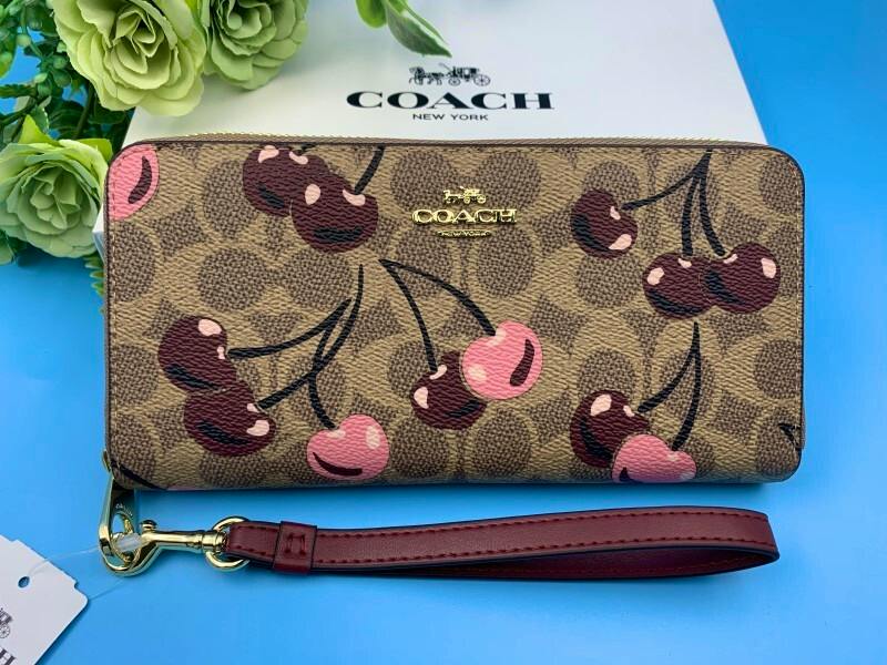 Front view of a Coach signature long wallet with cherry print and a detachable wrist strap on a blue background.