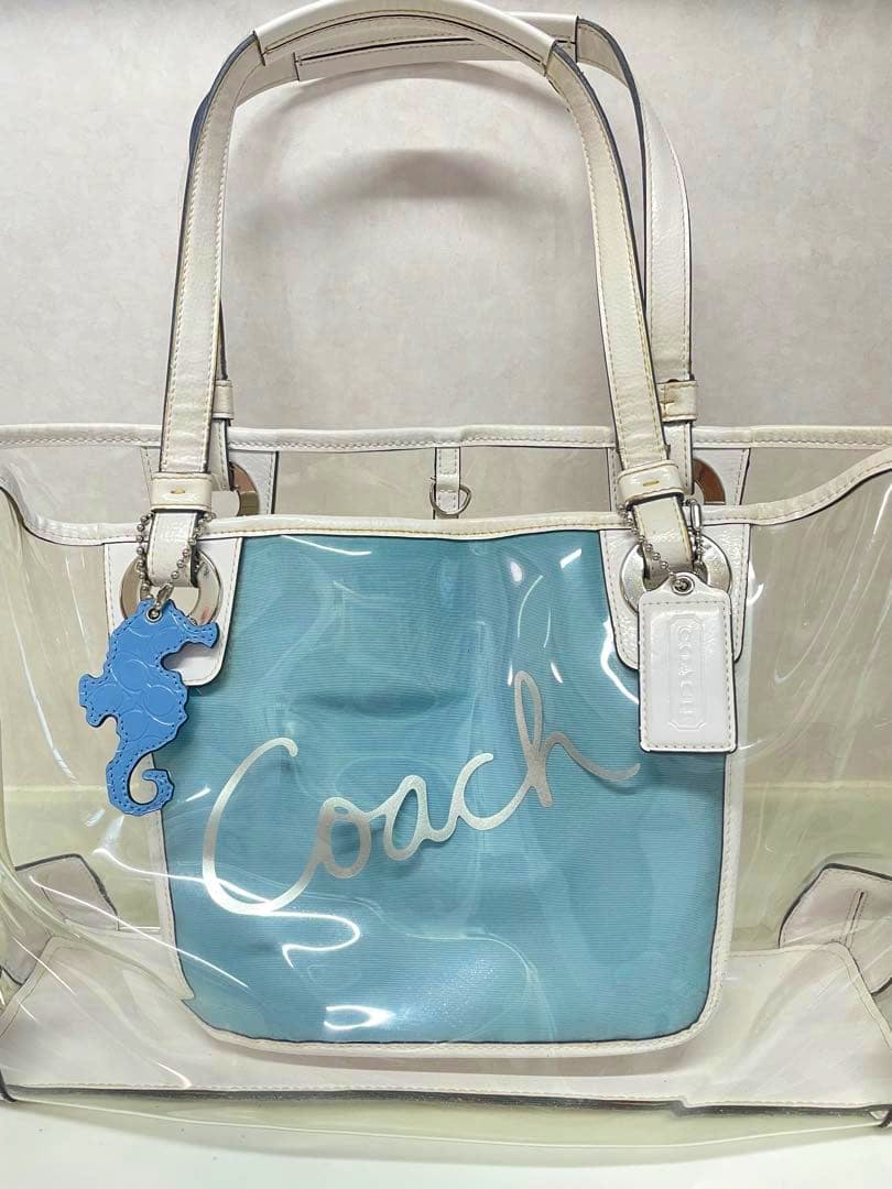 Clear Coach tote bag with white leather trim and handles, a blue inner pouch, and a blue seahorse charm attached.
