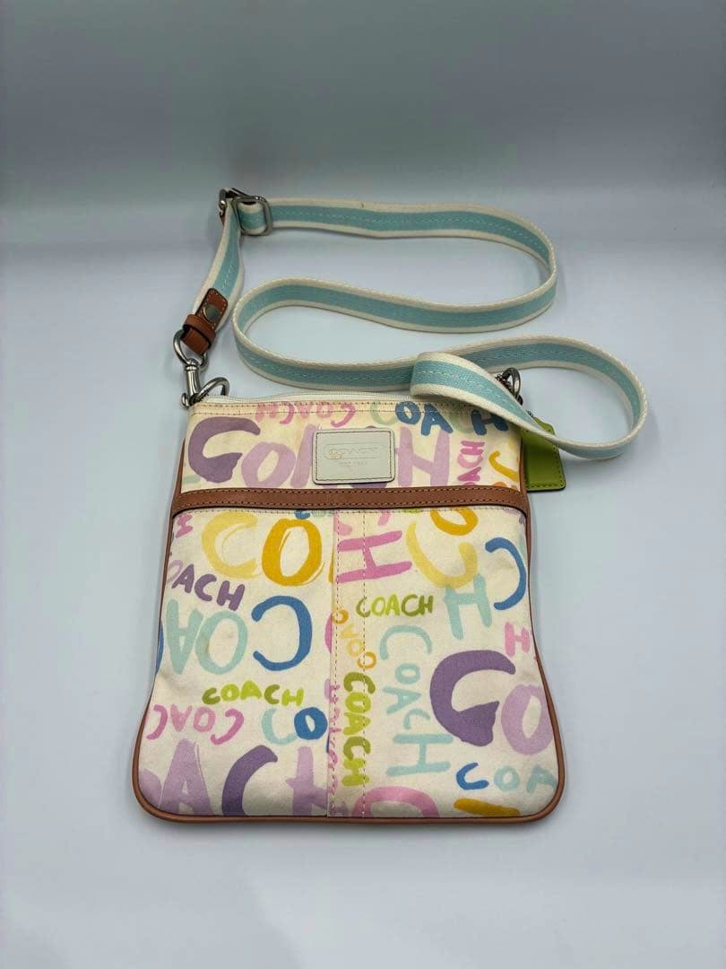 COACH canvas shoulder bag with colorful logo print, leather trim, and a light-blue striped strap photographed flat.