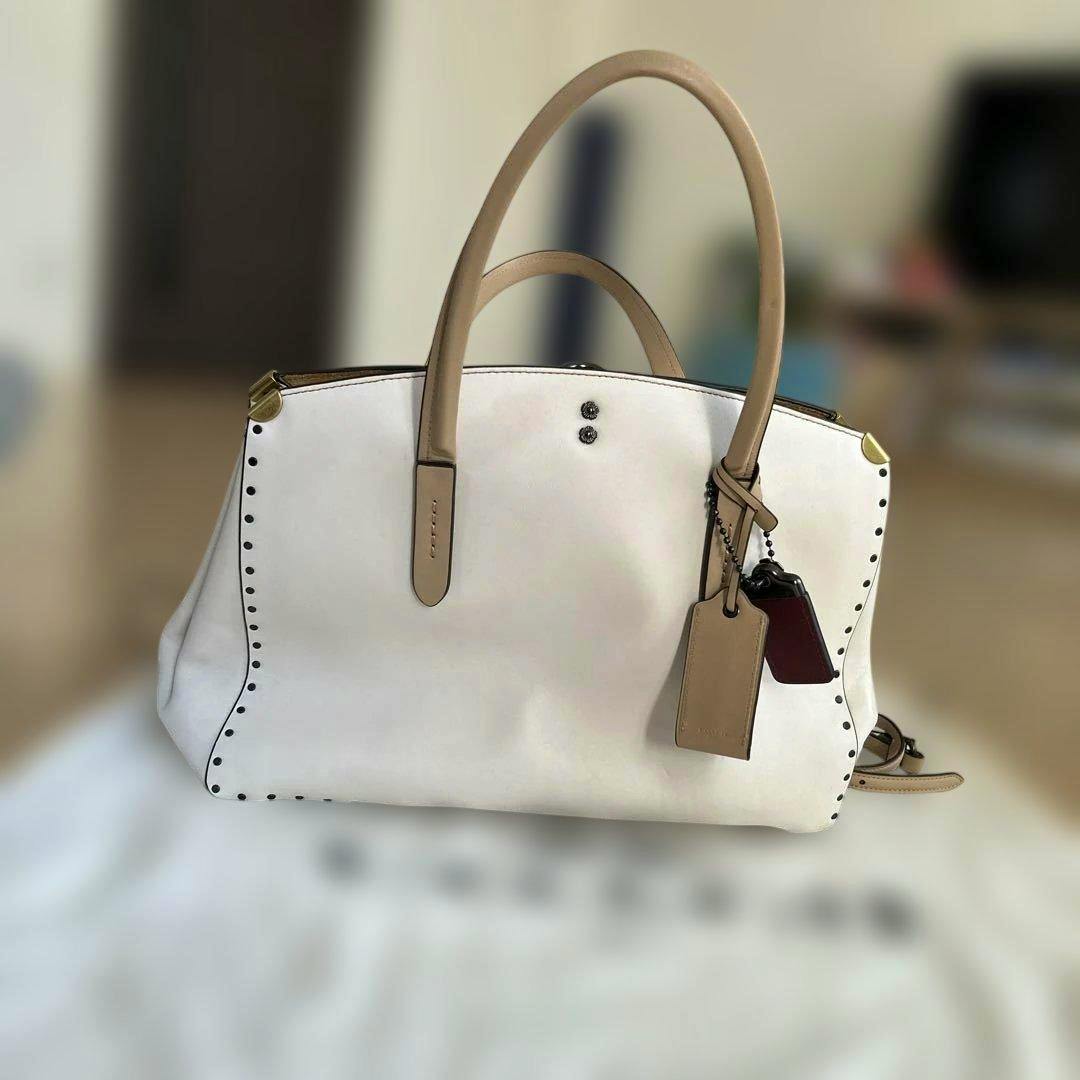 Front view of the Coach Cooper Carryall cow leather bag in white with tan handles, studded edges, and hanging tags.