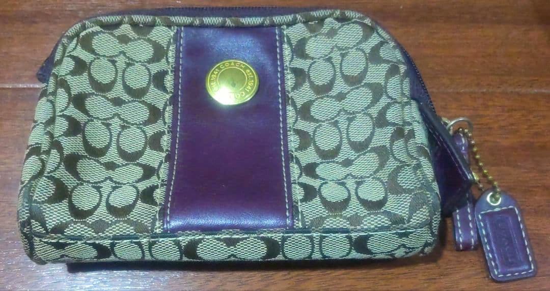 Front view of a Coach cotton pouch with signature 'C' patterned fabric, a central leather strip with a round metal Coach emblem, and a leather zipper pull.