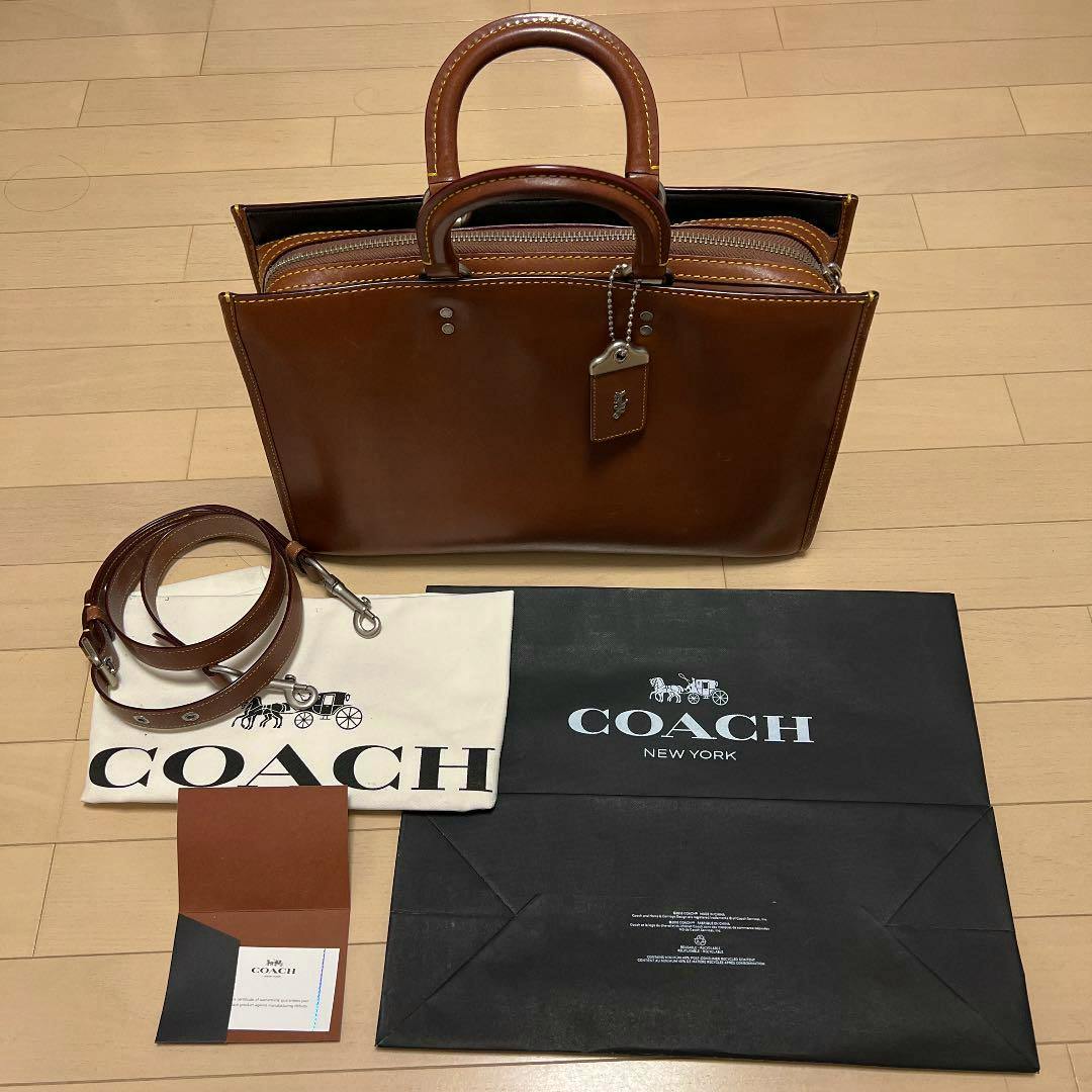 Brown cow-leather Coach bag with top handles shown with a detachable strap, dust bag, and paperwork on a wood floor.