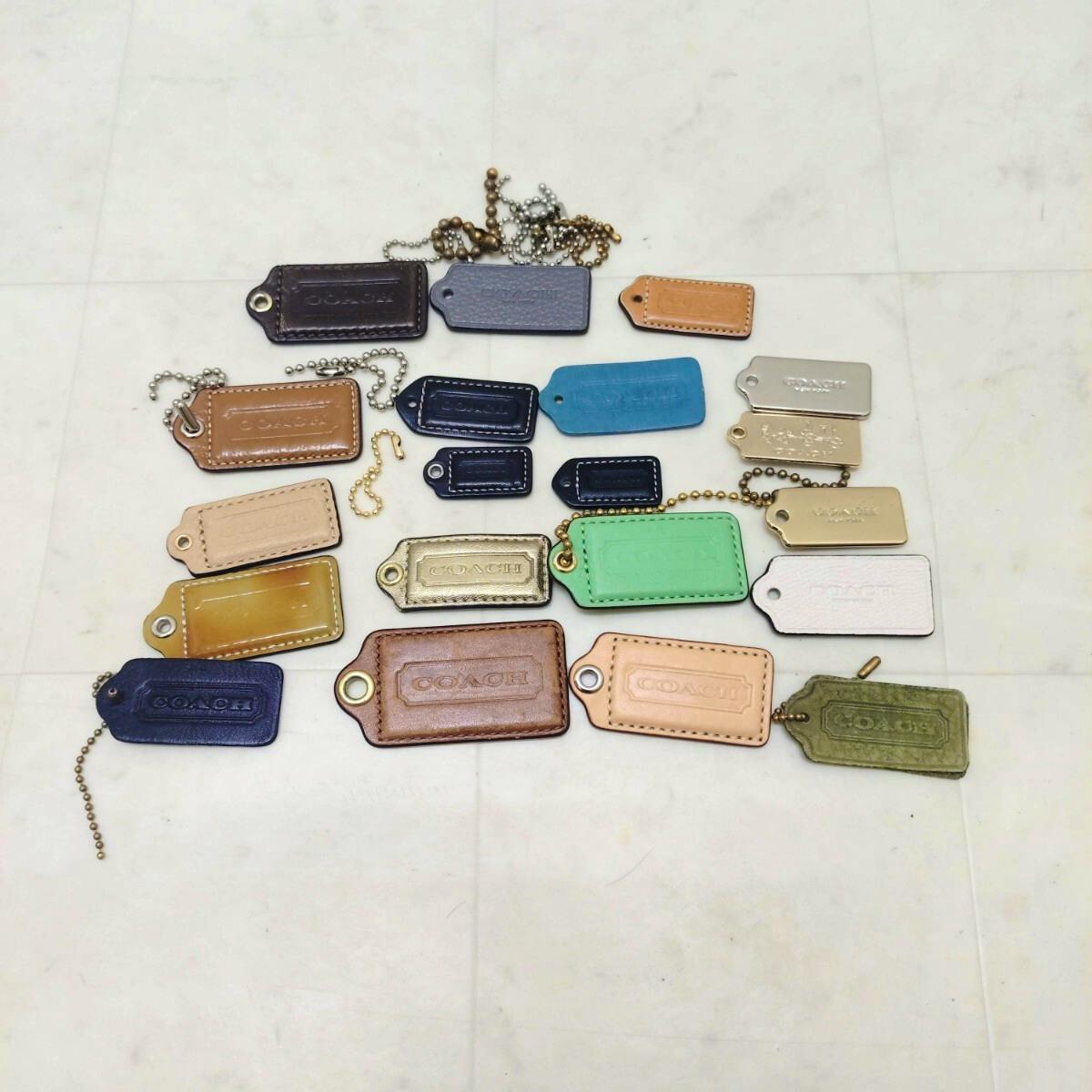 Multiple COACH leather and metal charm tags in various colors laid out on a white tiled surface.