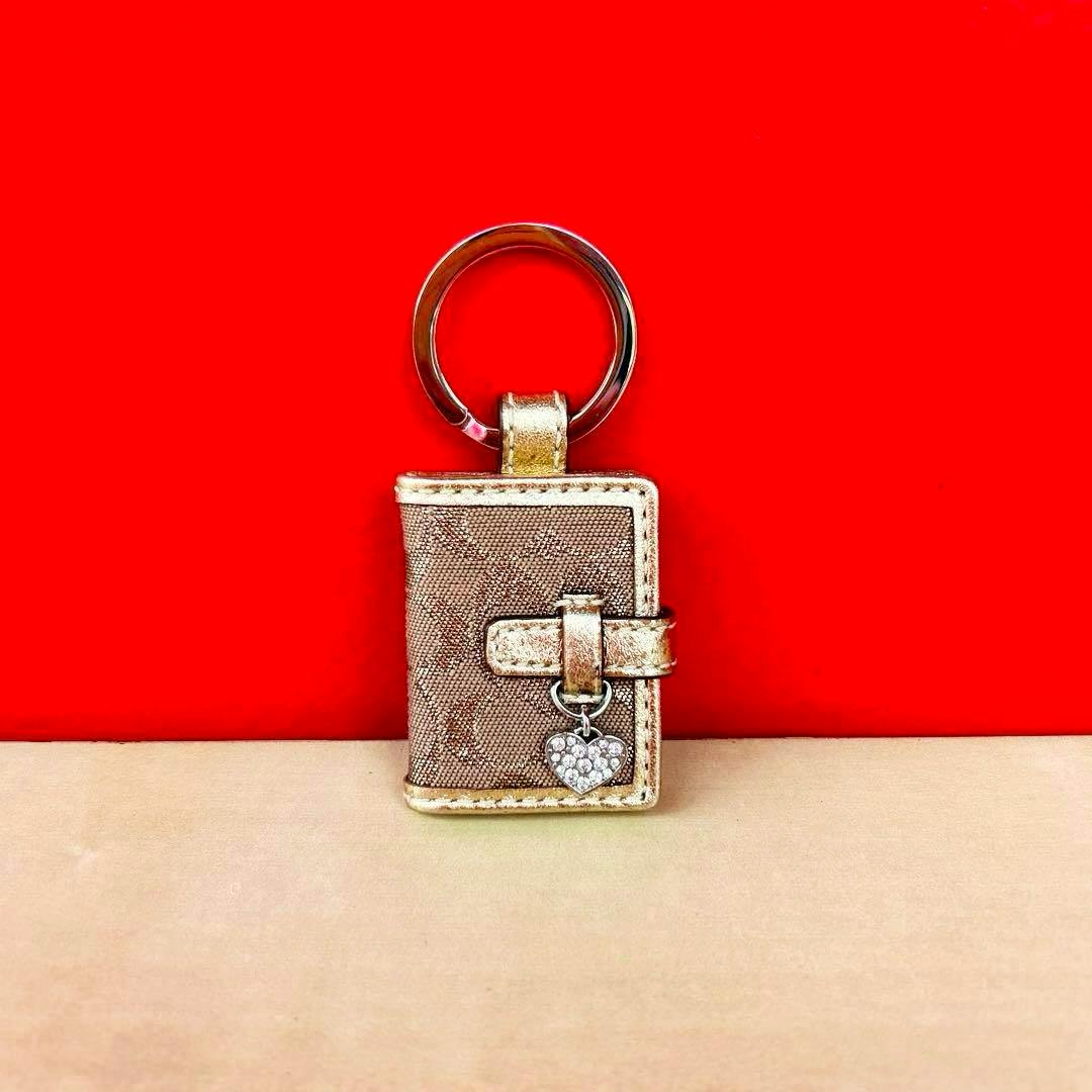 COACH cow leather keychain shaped like a small book with a heart charm on a metal key ring against a red background.