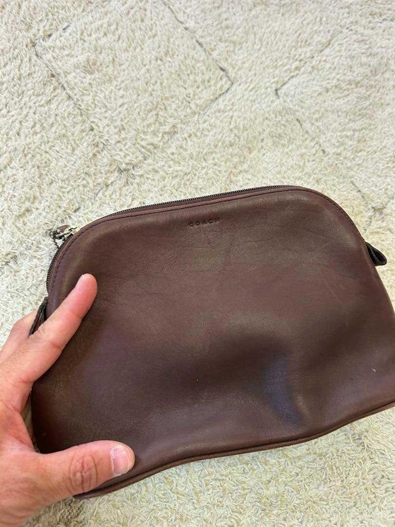 A brown COACH cow leather pouch held in a hand on a light textured rug.